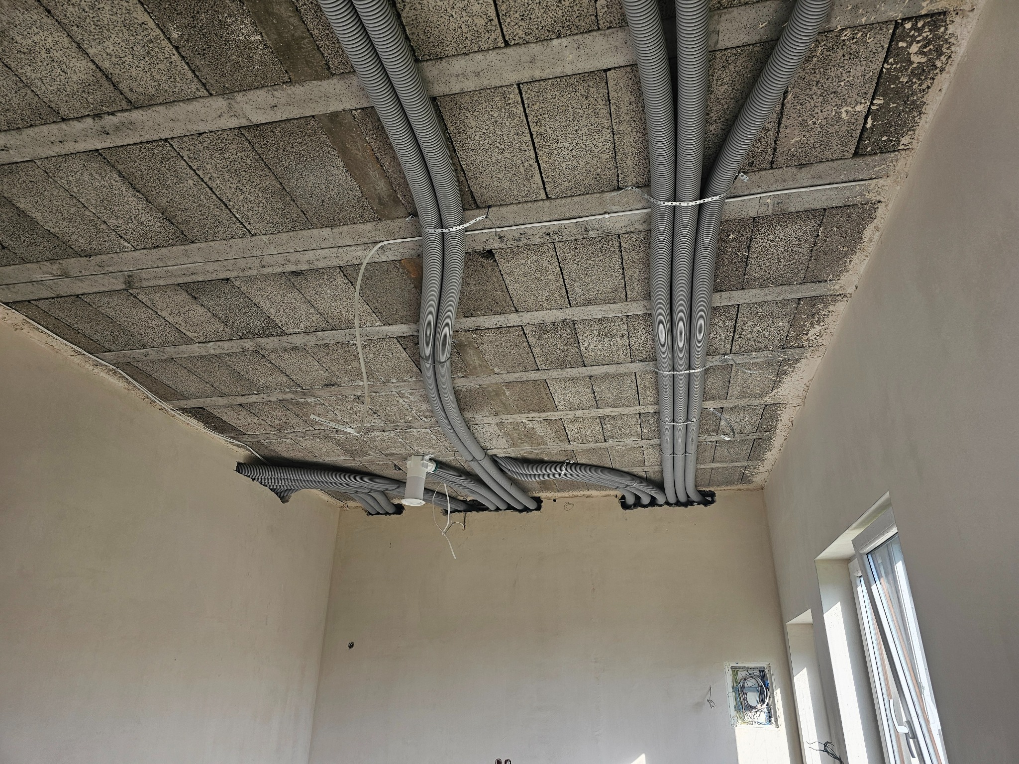 Szare rury instalacyjne przymocowane do sufitu z betonowych płyt w surowym wnętrzu. Widoczne otwory w ścianach, okno i wystające kable.
