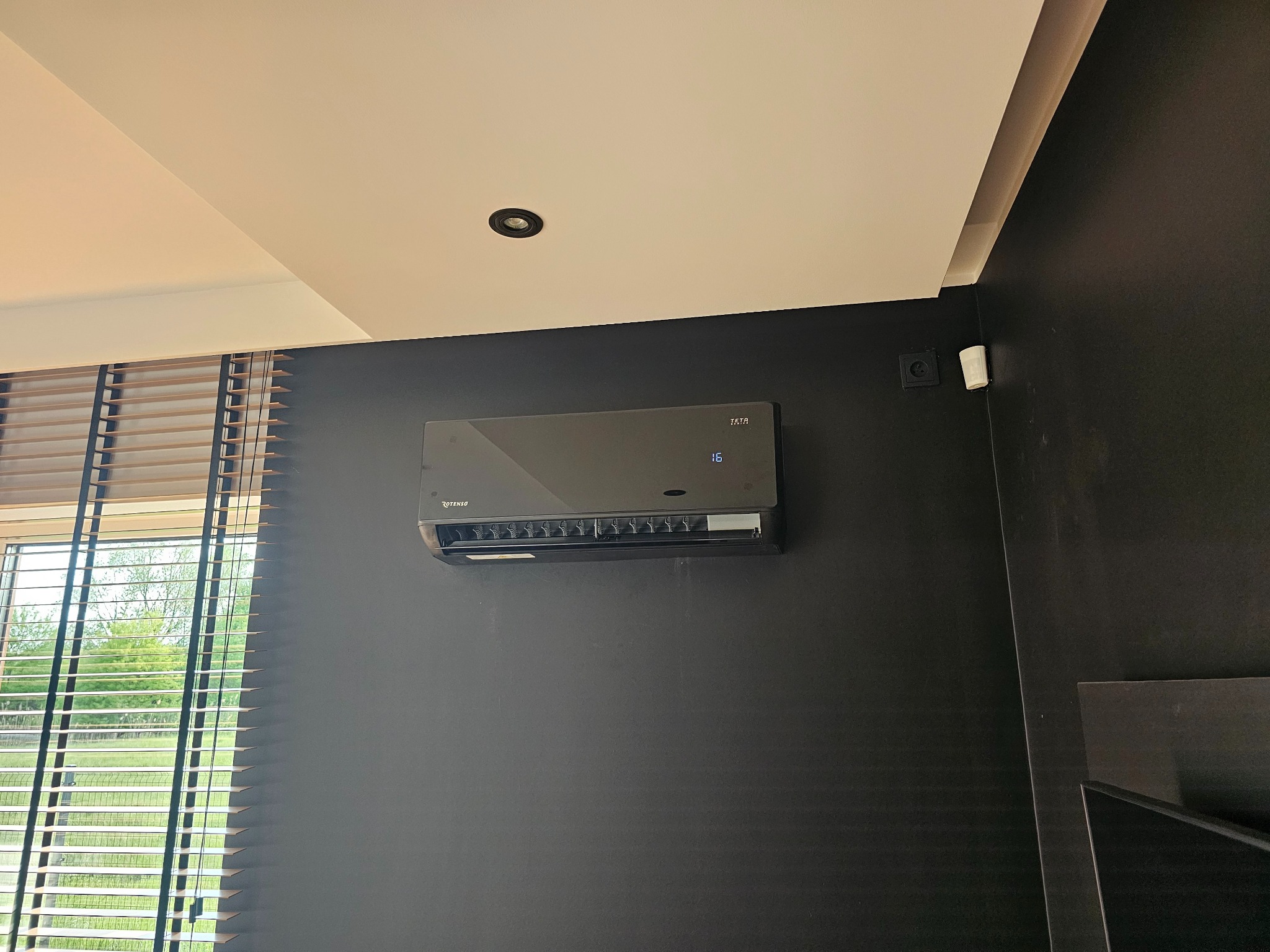 Czarny klimatyzator Rotenso z wyświetlaczem 16 stopni Celsjusza, zamontowany na ciemnej ścianie w nowoczesnym wnętrzu z żaluzjami w oknie.