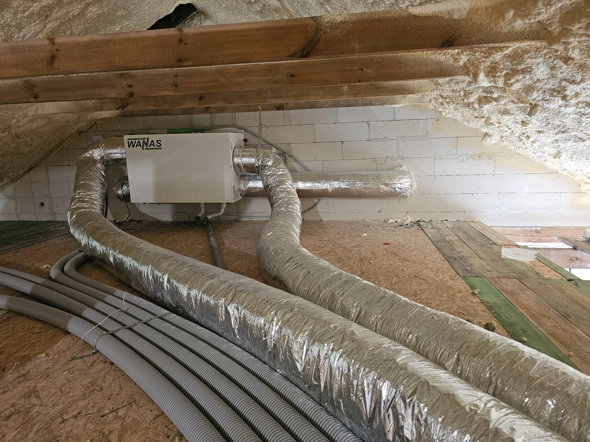 Biała centrala rekuperacyjna WANAS z podłączonymi przewodami wentylacyjnymi na poddaszu, widoczne ocieplenie pianą PUR i bloczki z betonu komórkowego.