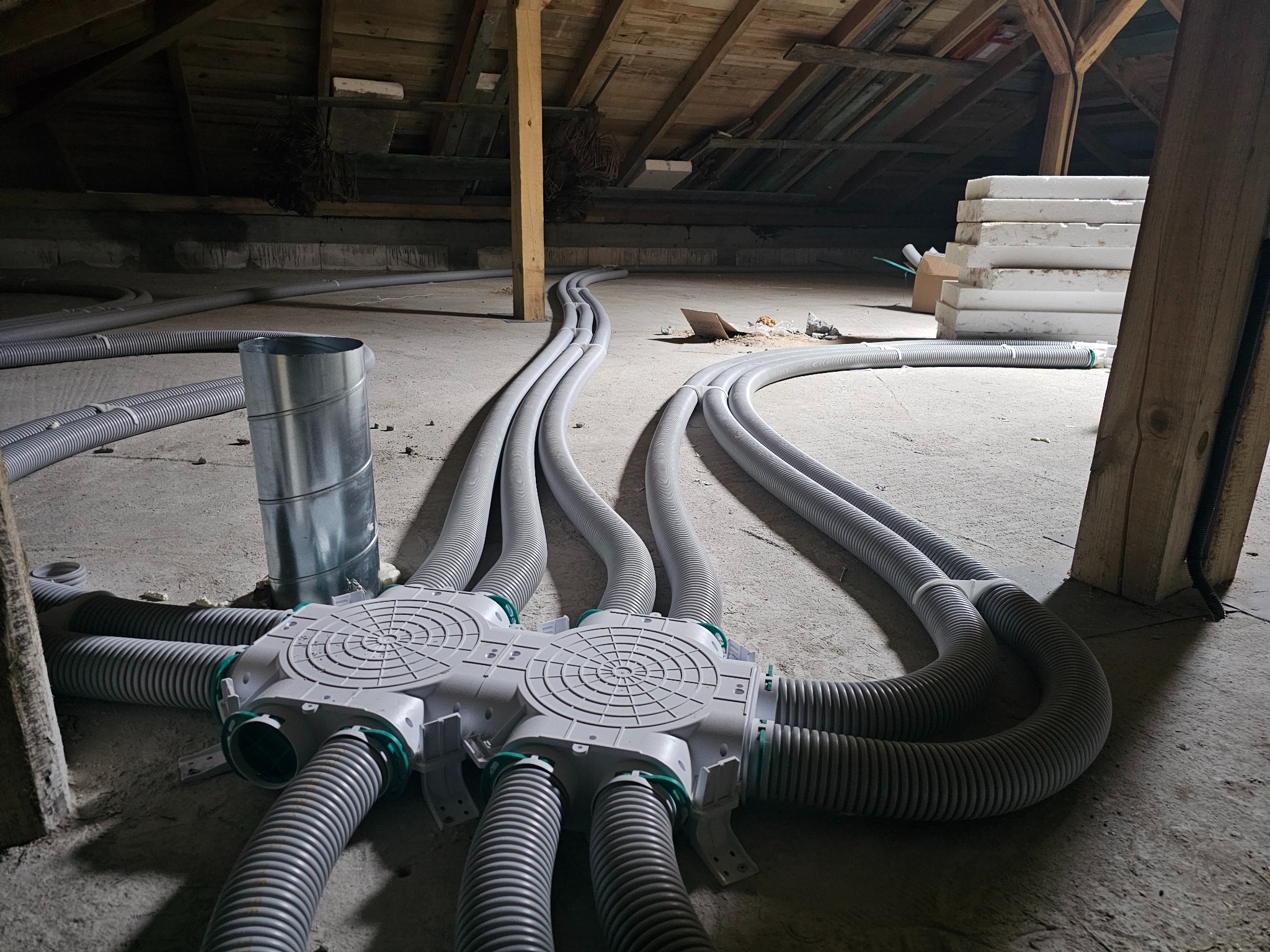 Szare, karbowane rury wentylacyjne rozchodzą się z białej skrzynki rozdzielczej na poddaszu. Widoczne drewniane elementy konstrukcji dachu i izolacja.
