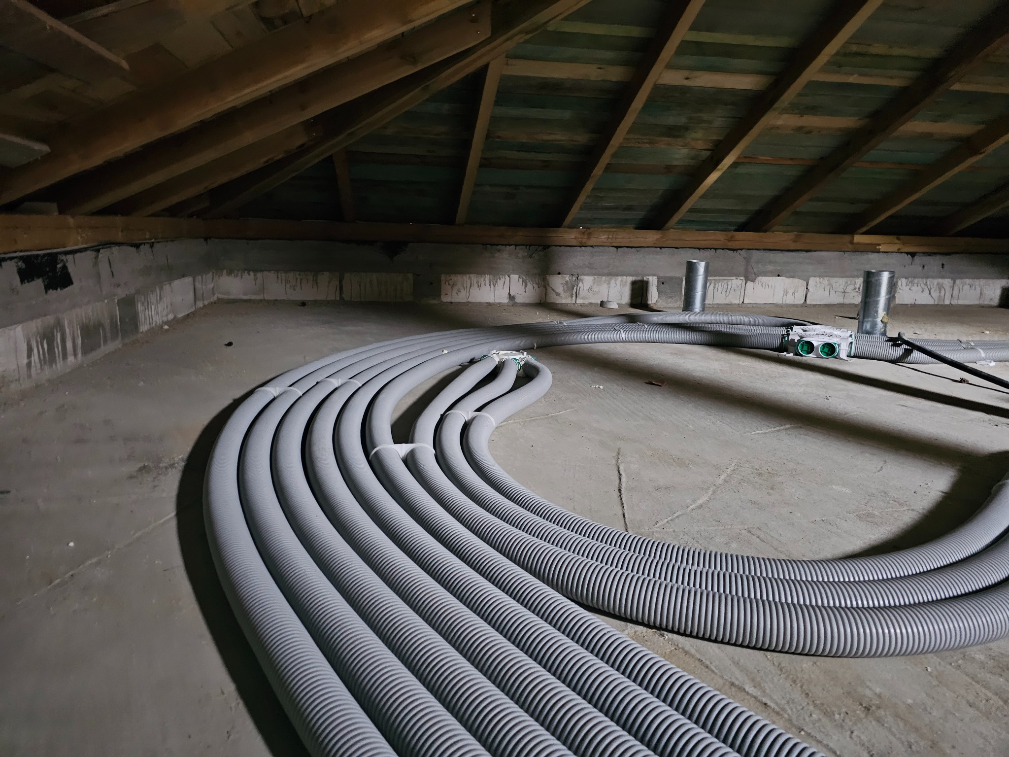 Szare, karbowane rury wentylacyjne ułożone równolegle na poddaszu z drewnianą konstrukcją dachu. Widoczne elementy instalacji i betonowa podłoga.
