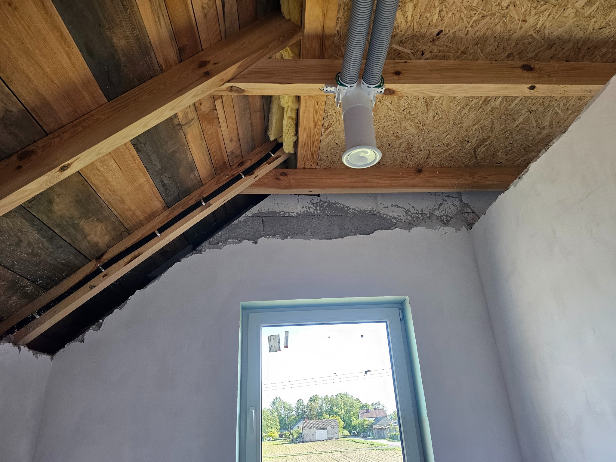 Montaż rekuperacji na poddaszu: widoczne rury wentylacyjne, centrala rekuperacyjna pod stropem z desek, okno z widokiem na pole i zabudowania.