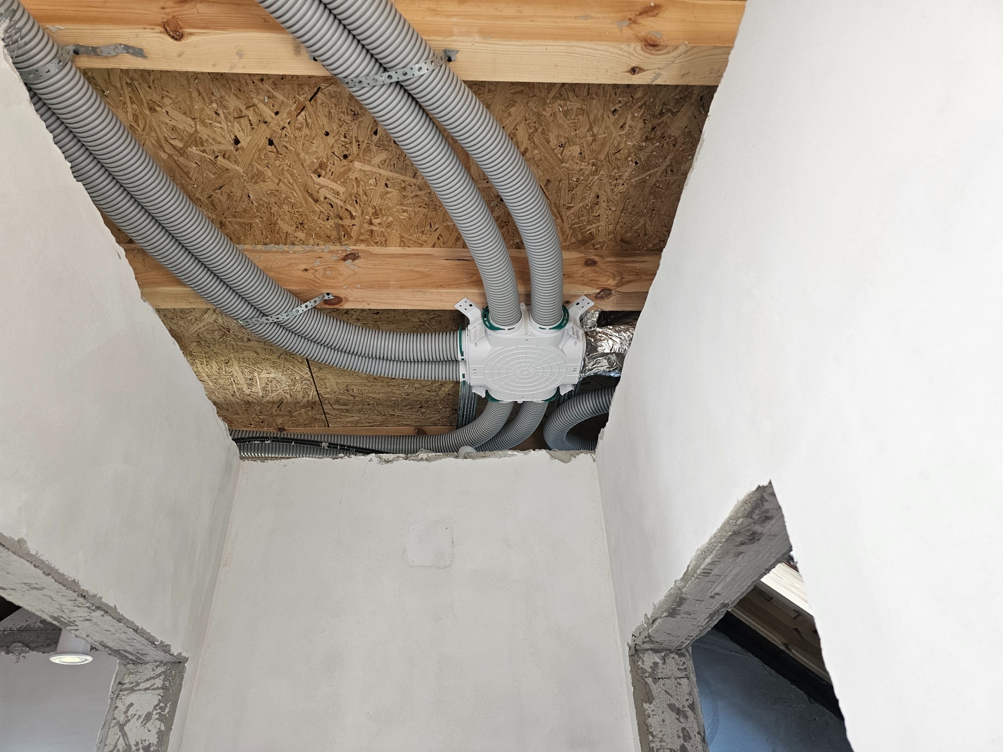 Szare, karbowane rury wentylacyjne przymocowane do drewnianego stropu, wchodzące do białej skrzynki rozdzielczej w surowym wnętrzu budynku.