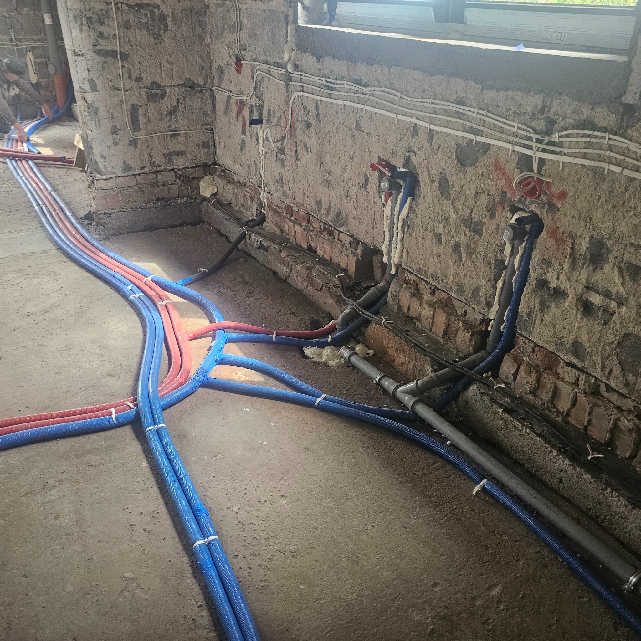 Instalacja rur grzewczych (czerwone i niebieskie) oraz kanalizacyjnych (szare) w surowym wnętrzu z ceglanymi ścianami. Widoczne łączenia i mocowania.