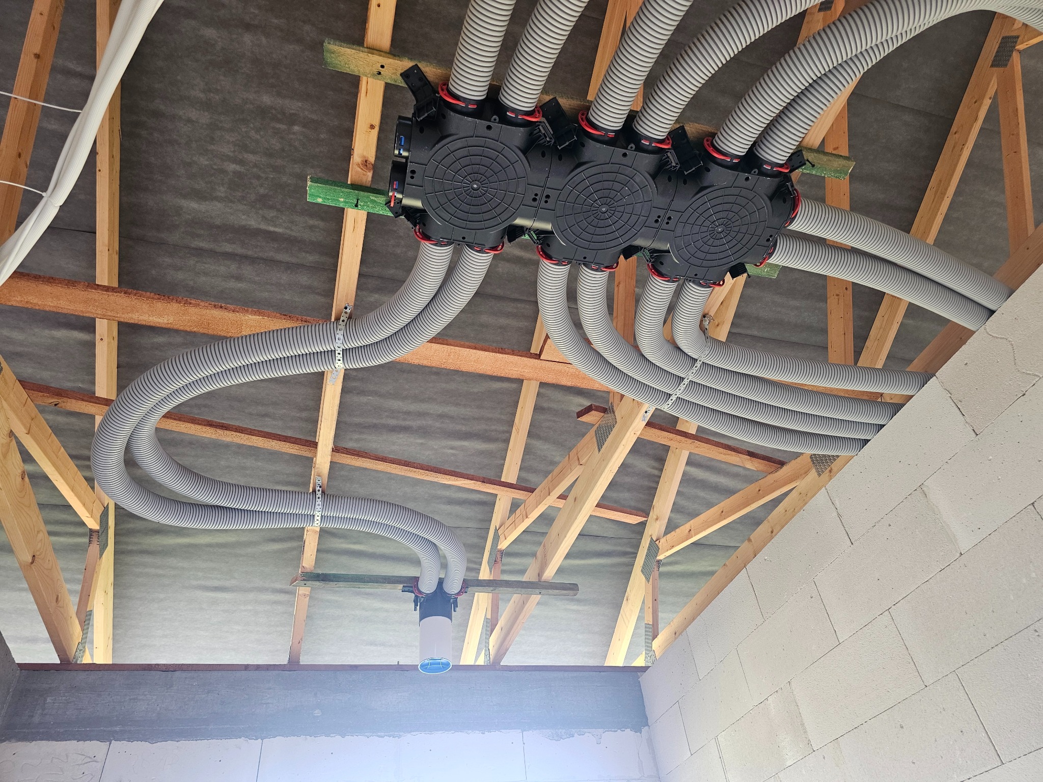 Rozprowadzenie szarych, karbowanych rur wentylacyjnych pod stropem, przymocowanych do czarnej skrzynki rozdzielczej na drewnianej konstrukcji. Widok z dołu na instalację.