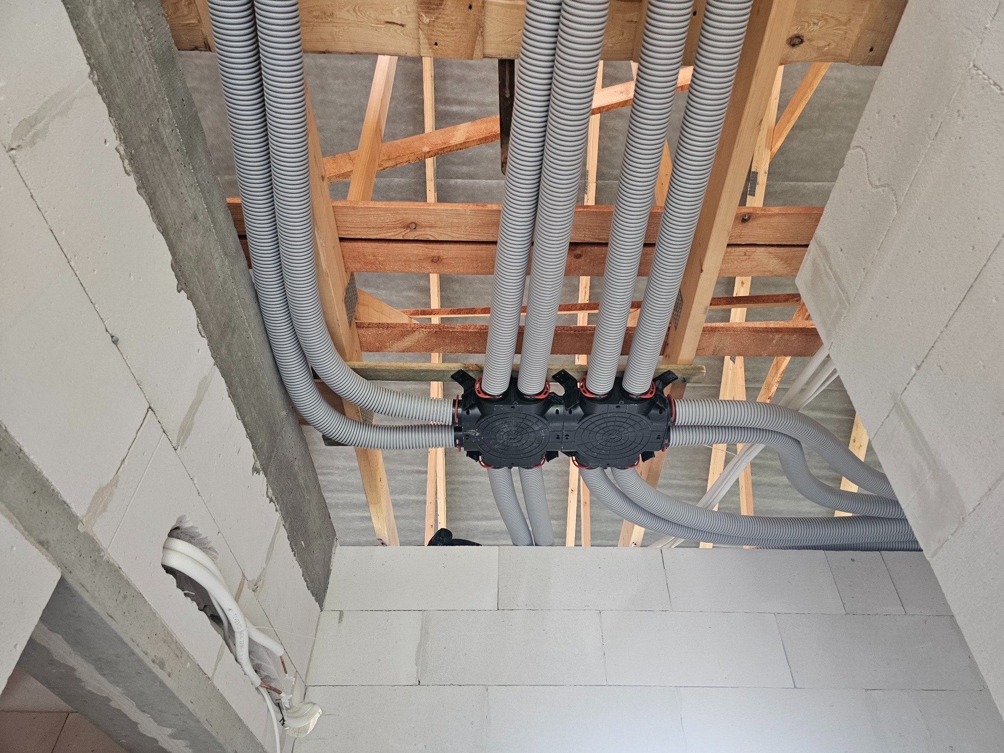 Szare, karbowane rury wentylacyjne prowadzone przez drewnianą konstrukcję stropu, połączone z czarną skrzynką rozdzielczą w surowym wnętrzu budynku z białych bloczków.