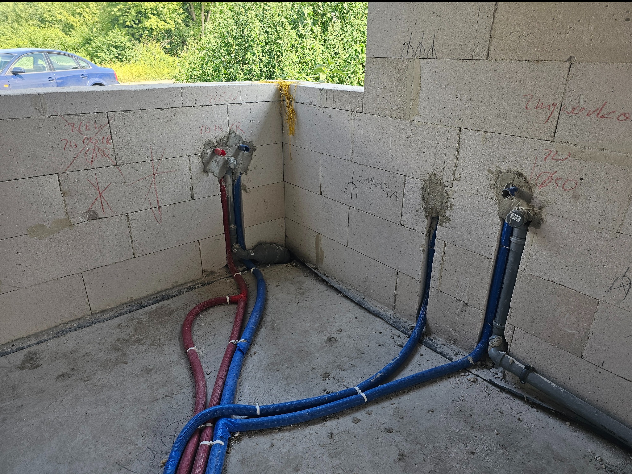 Instalacja wodna w surowym wnętrzu: czerwone i niebieskie rury prowadzone wzdłuż ściany z betonu komórkowego, z widocznymi oznaczeniami i elementami montażowymi.