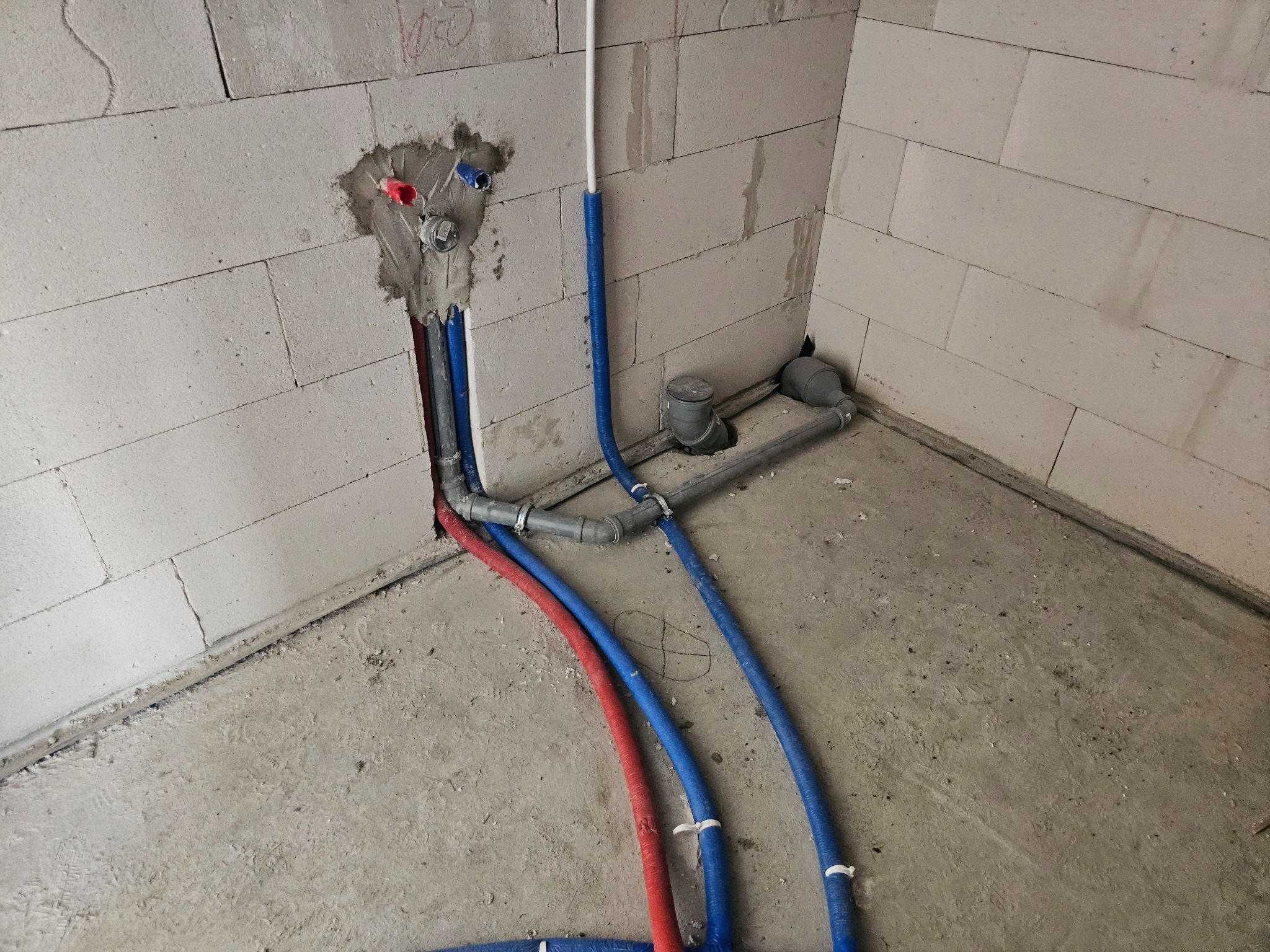 Instalacja wodno-kanalizacyjna w surowym wnętrzu: czerwone i niebieskie rury prowadzone wzdłuż ściany z betonu komórkowego, szare rury odpływowe.