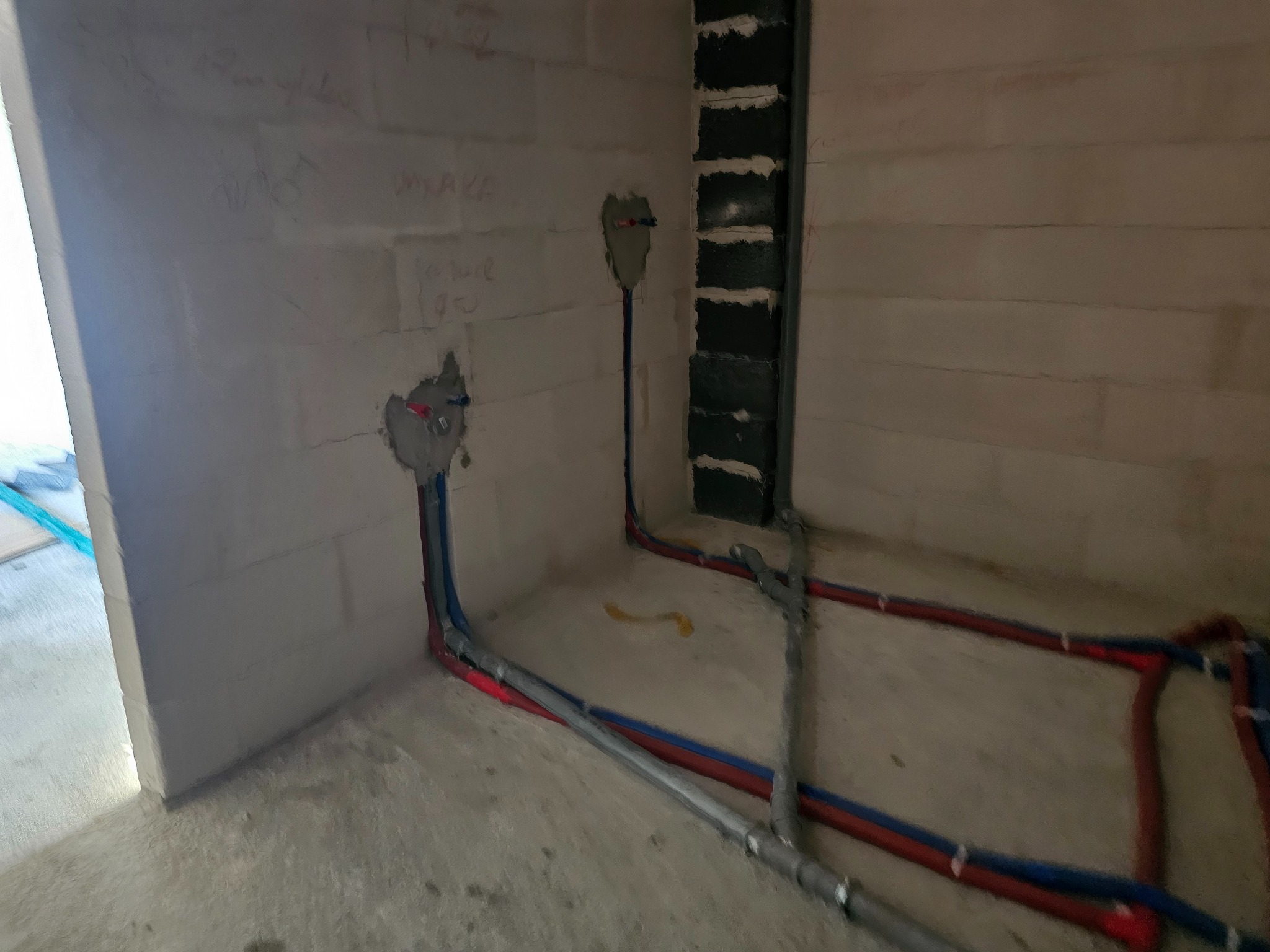 Surowe wnętrze z instalacją wodno-kanalizacyjną w trakcie montażu. Widoczne rury w kolorach niebieskim i czerwonym, prowadzone wzdłuż ścian z betonu komórkowego.