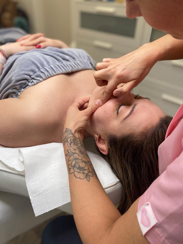 Kobieta poddawana masażowi twarzy w gabinecie kosmetycznym; widoczne dłonie masażystki, fragment tatuażu na jej ramieniu oraz zamknięte oczy klientki leżącej na białym podkładzie.