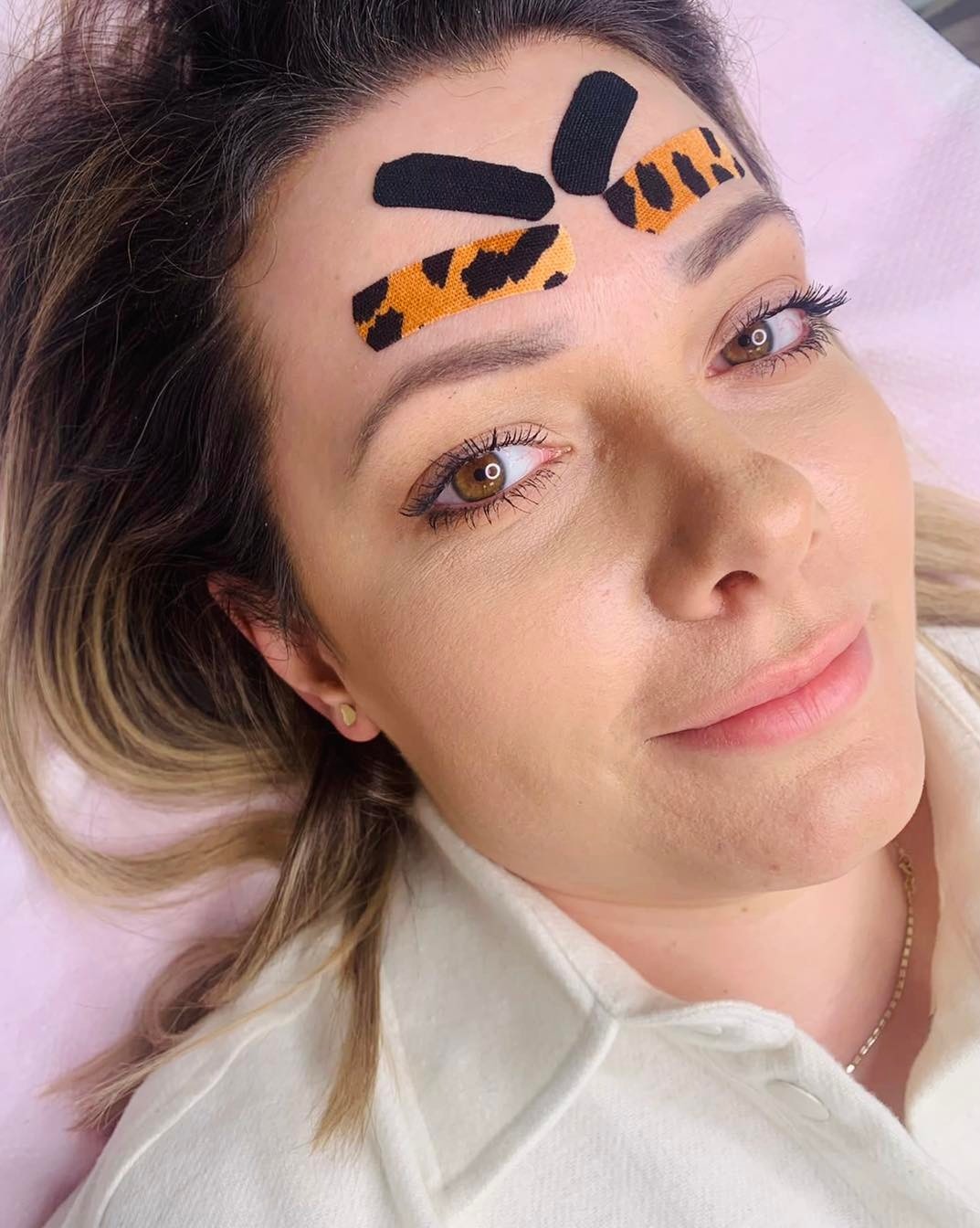 Kobieta po zabiegu tapingu twarzy z aplikacjami w czarne i panterkowe wzory na czole, leżąca na różowym podłożu.