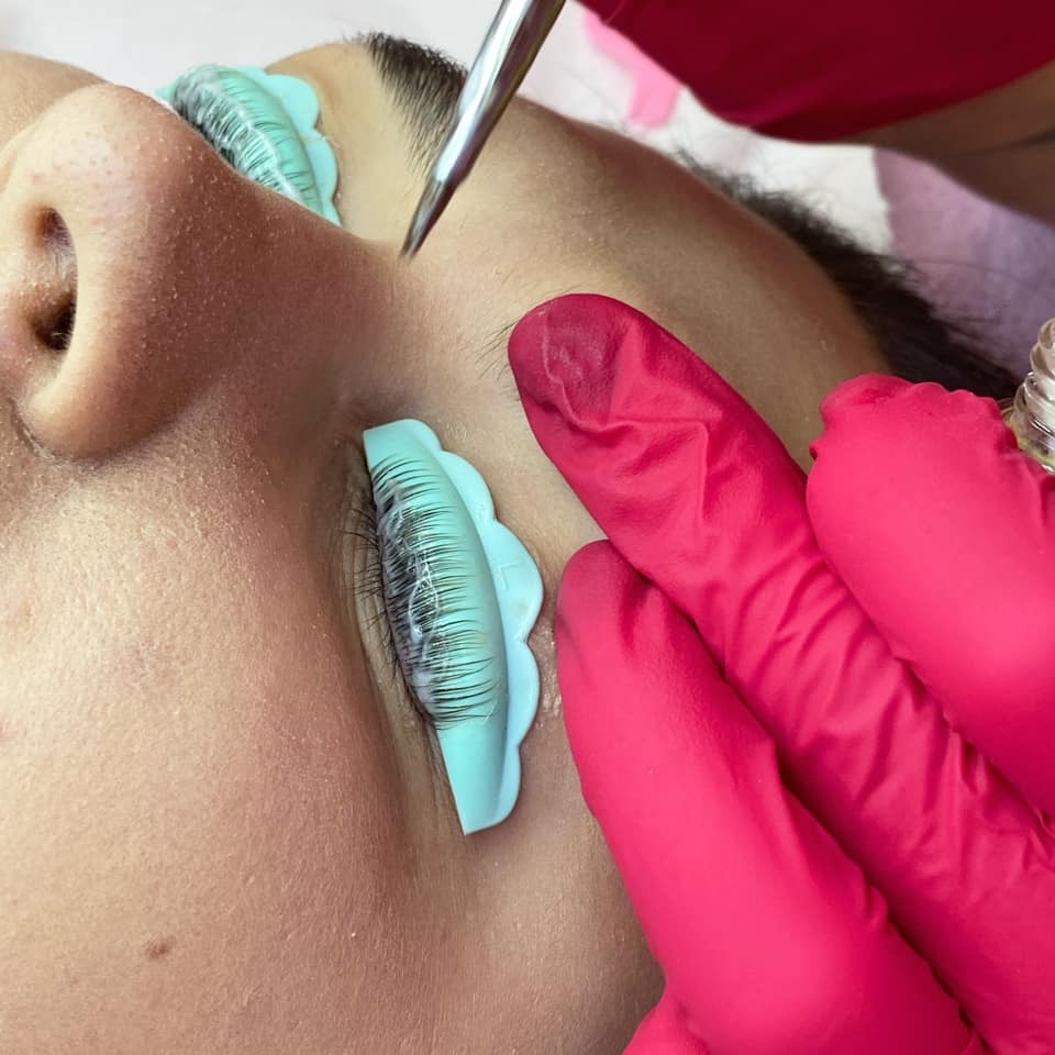 Zabieg liftingu rzęs: rzęsy przyklejone do silikonowej formy, pęseta i dłoń w różowej rękawiczce aplikują preparat.