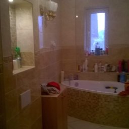 Kamil Wilczek - Wykończona łazienka z beżowymi płytkami, mozaiką wokół wanny, toaletą z czerwonym ręcznikiem, szafką pod umywalką i oknem z widokiem na zewnątrz.