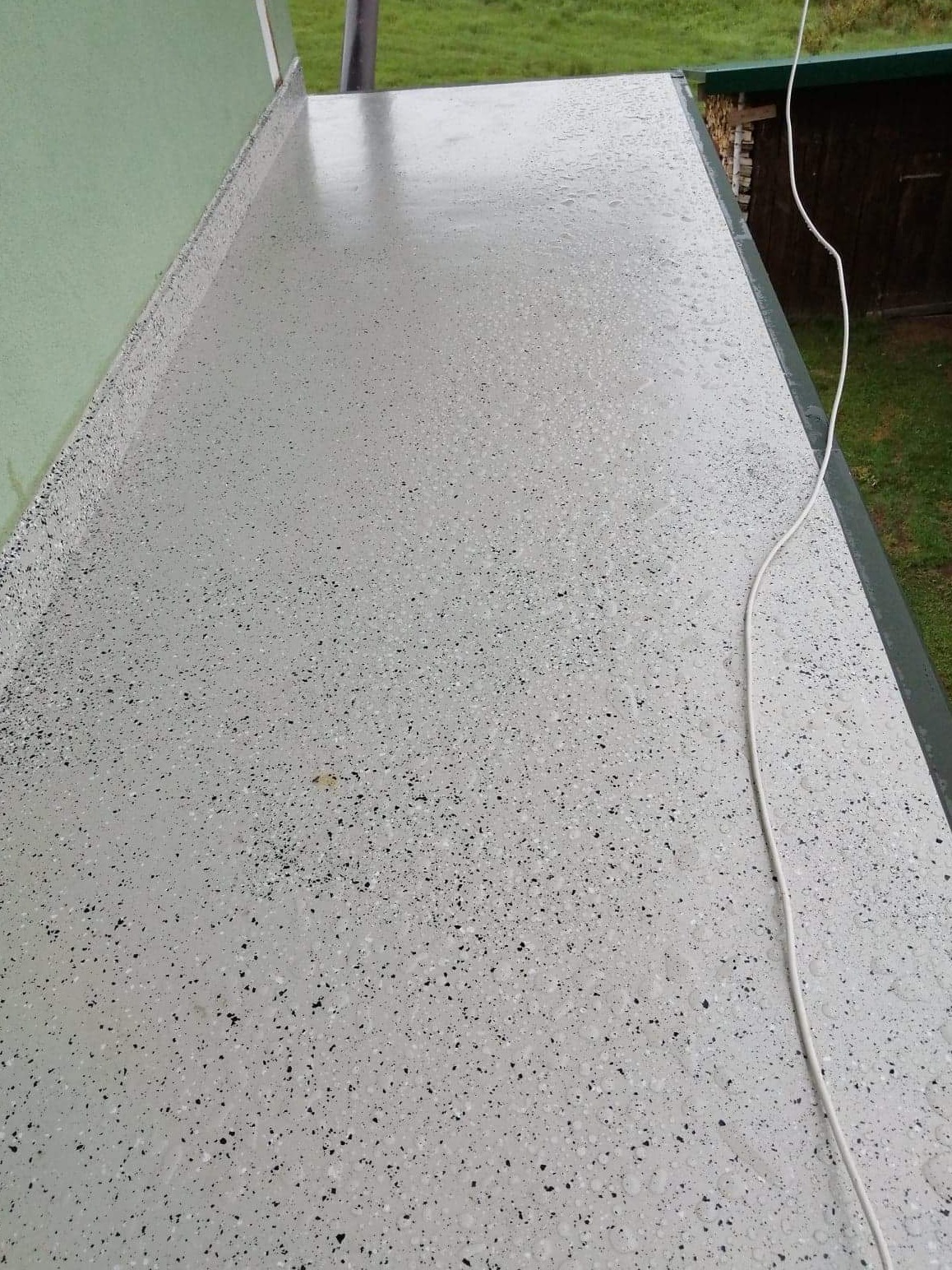 Świeżo położona, jasnoszara, żywiczna posadzka balkonowa z drobnymi, czarnymi plamkami, widoczna z góry, z fragmentem zielonej ściany i kablem elektrycznym na krawędzi.