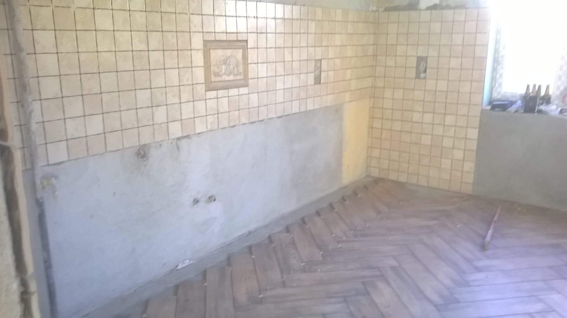 Wnętrze w trakcie prac remontowych: częściowo położone płytki ścienne, gładź na dolnej części ściany, drewniana podłoga ułożona w jodełkę, widoczne puste otwory po gniazdkach elektrycznych.