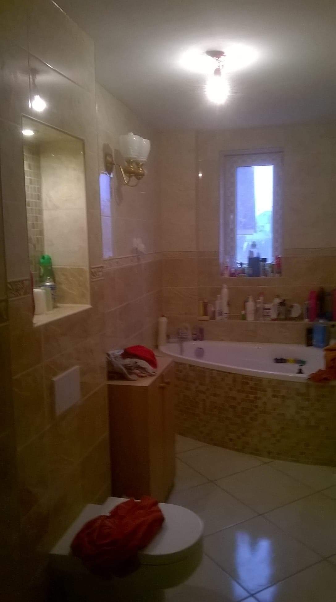 Wykończona łazienka z beżowymi płytkami, mozaiką wokół wanny, toaletą z czerwonym ręcznikiem, szafką pod umywalką i oknem z widokiem na zewnątrz.