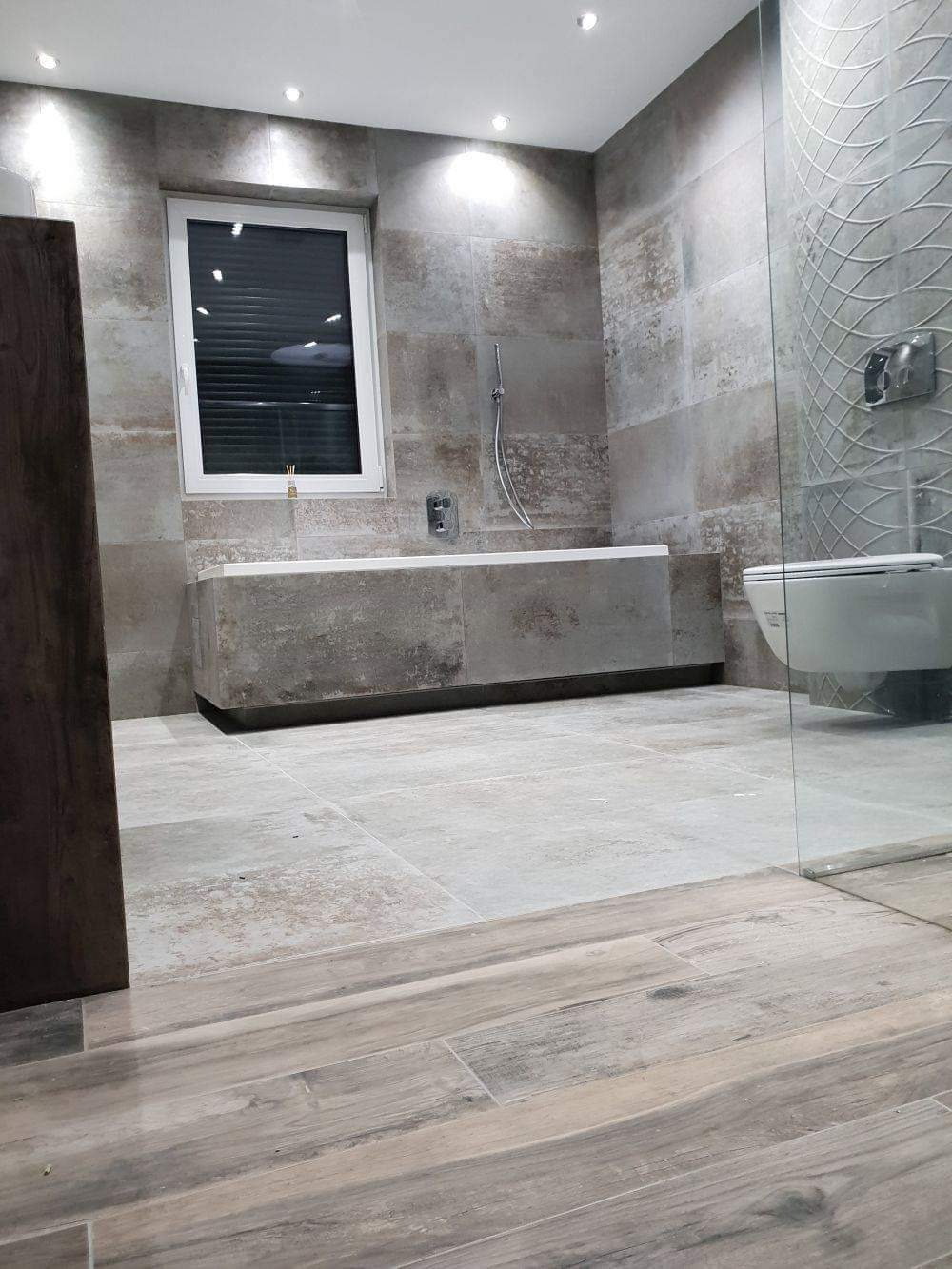 Nowoczesna łazienka z szarymi płytkami imitującymi kamień na ścianach i podłodze, z zabudowaną wanną, białą toaletą za szklaną ścianką kabiny prysznicowej i oknem z żaluzjami.