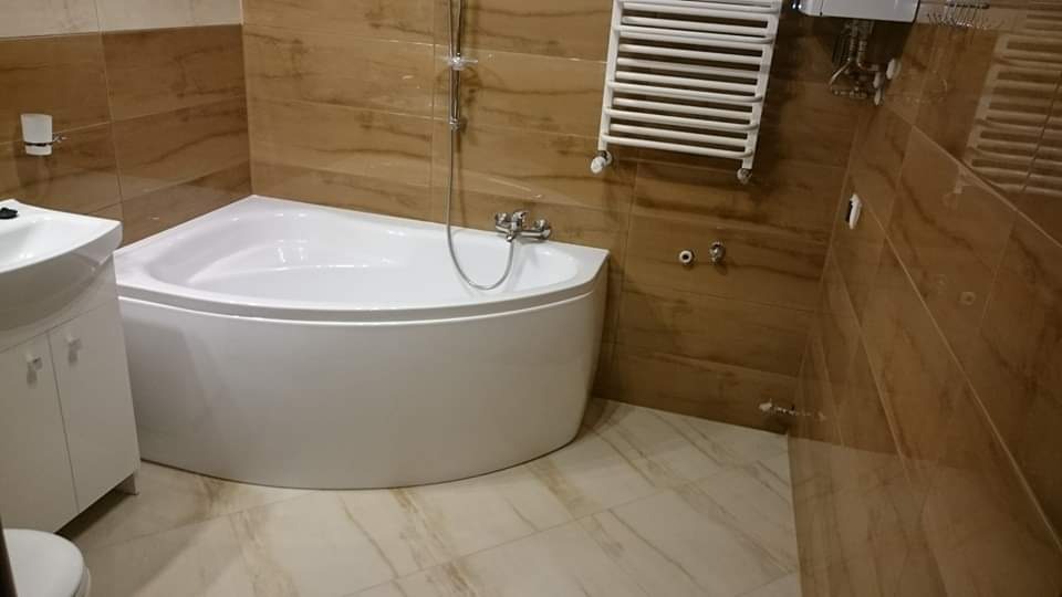 Narożna wanna z hydromasażem w jasnej łazience z beżowymi płytkami imitującymi drewno, grzejnik drabinkowy na ścianie, umywalka z szafką.