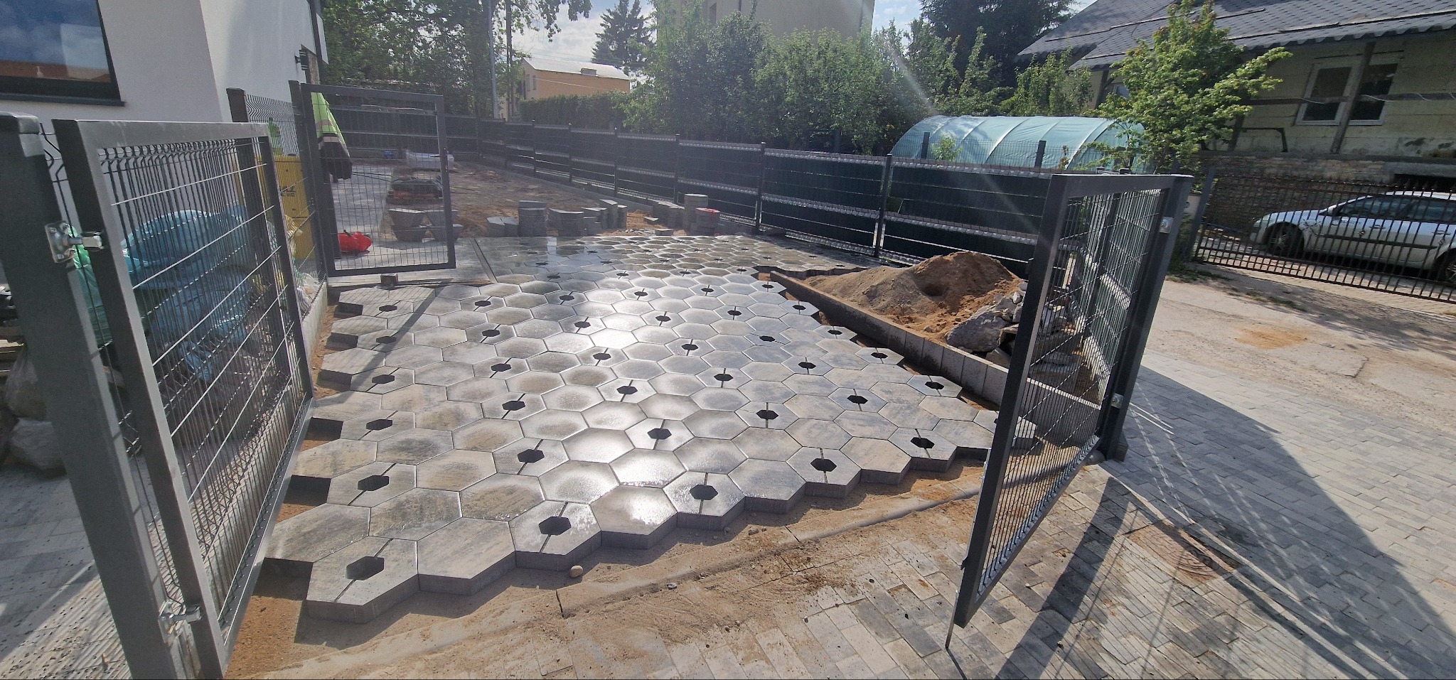 Układanie heksagonalnej kostki brukowej na posesji, widok zza metalowej bramy. Częściowo ułożona nawierzchnia, przygotowane podłoże, w tle dom i zieleń.