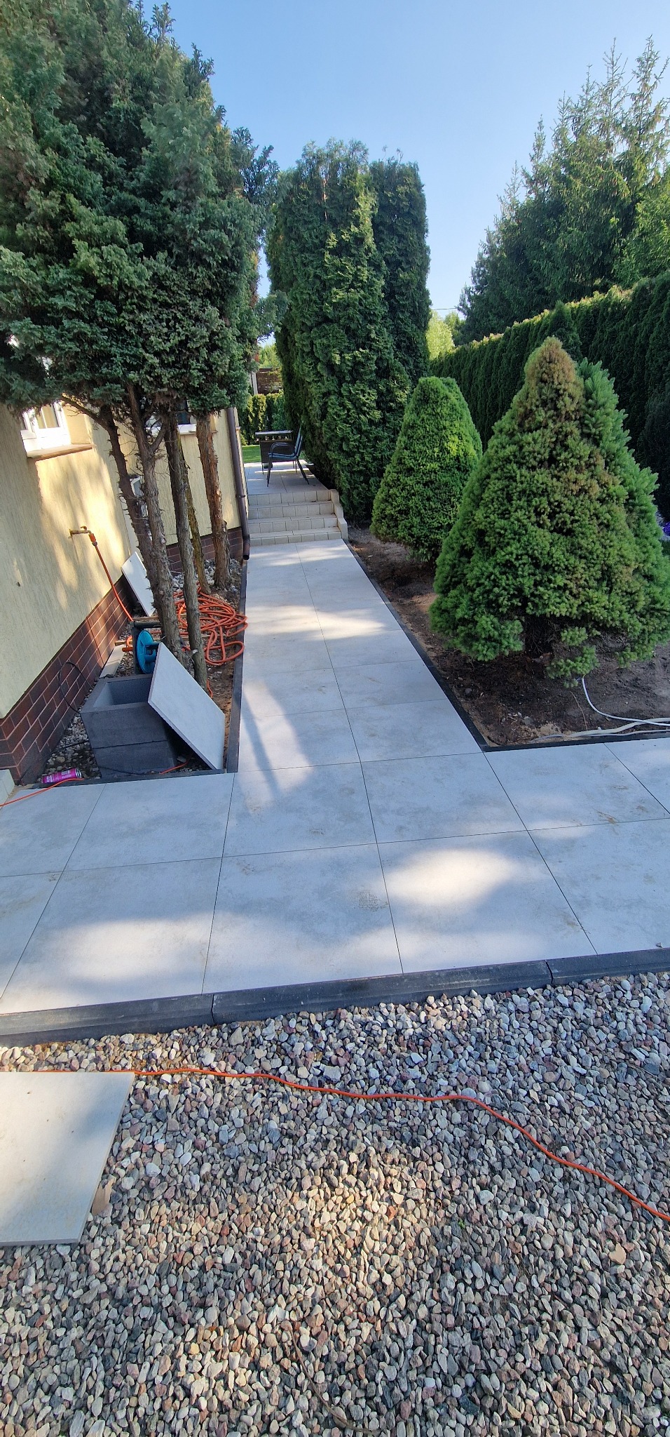 Jasnoszara ścieżka z płyt, obrzeża z kostki, ozdobne kamienie, iglaki. Widok na schody i meble ogrodowe w tle. Częściowo widoczne narzędzia i płyty.