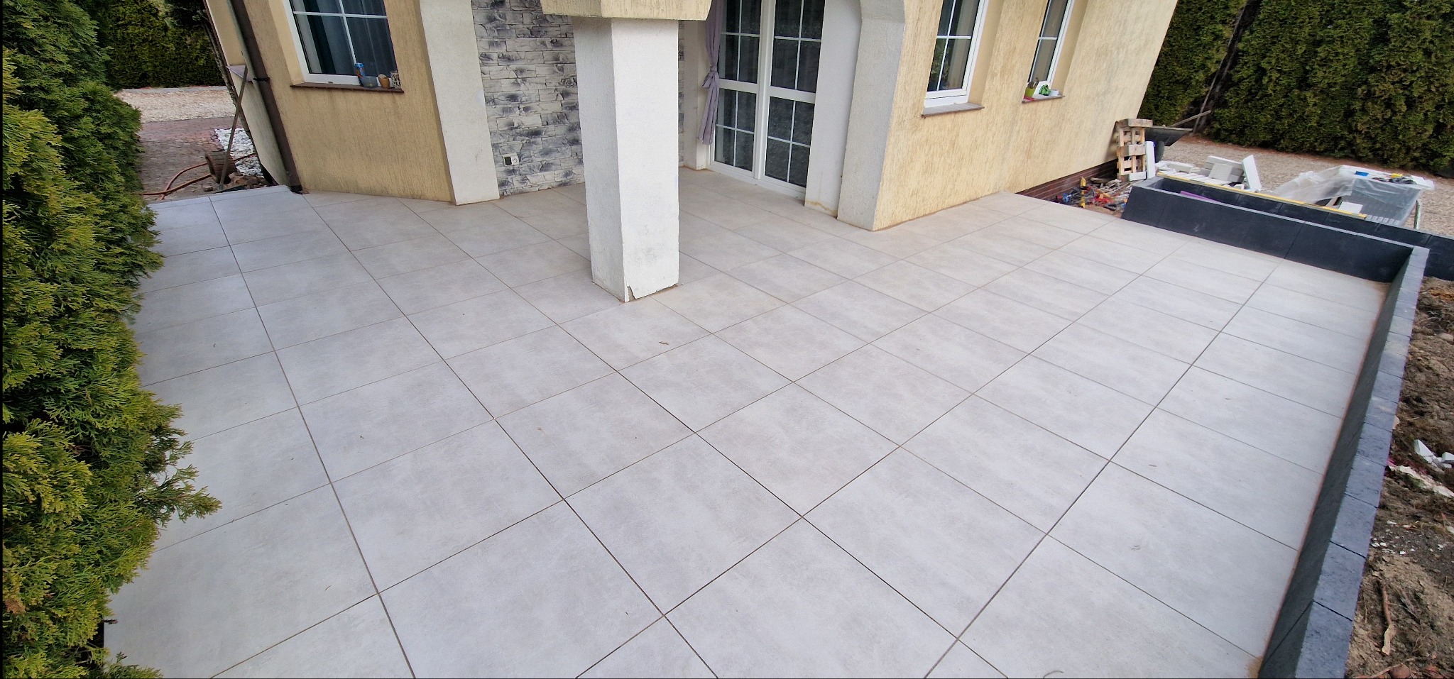 Jasnoszary taras z płytek ceramicznych przy domu z kolumną, widok z góry. W tle widoczne narzędzia i materiały budowlane.