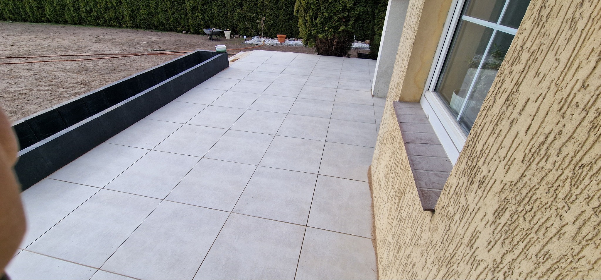 Jasne płytki tarasowe ułożone na zewnątrz budynku z oknem i czarną donicą. Widoczny fragment ogrodu z zielonym żywopłotem w tle. Minimalistyczny design.