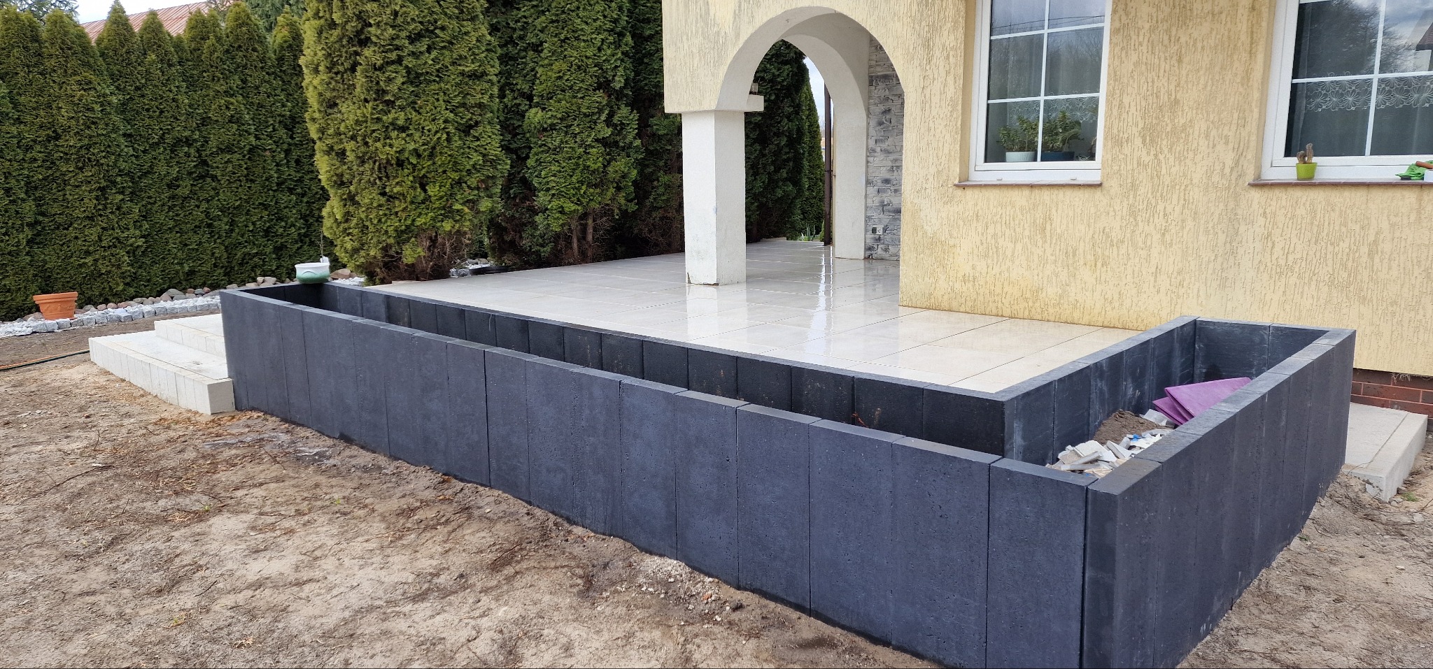 Nowoczesny taras z jasnymi płytkami i ciemną obudową z betonowych elementów. Widoczne schody wejściowe i fragment elewacji domu z oknem.