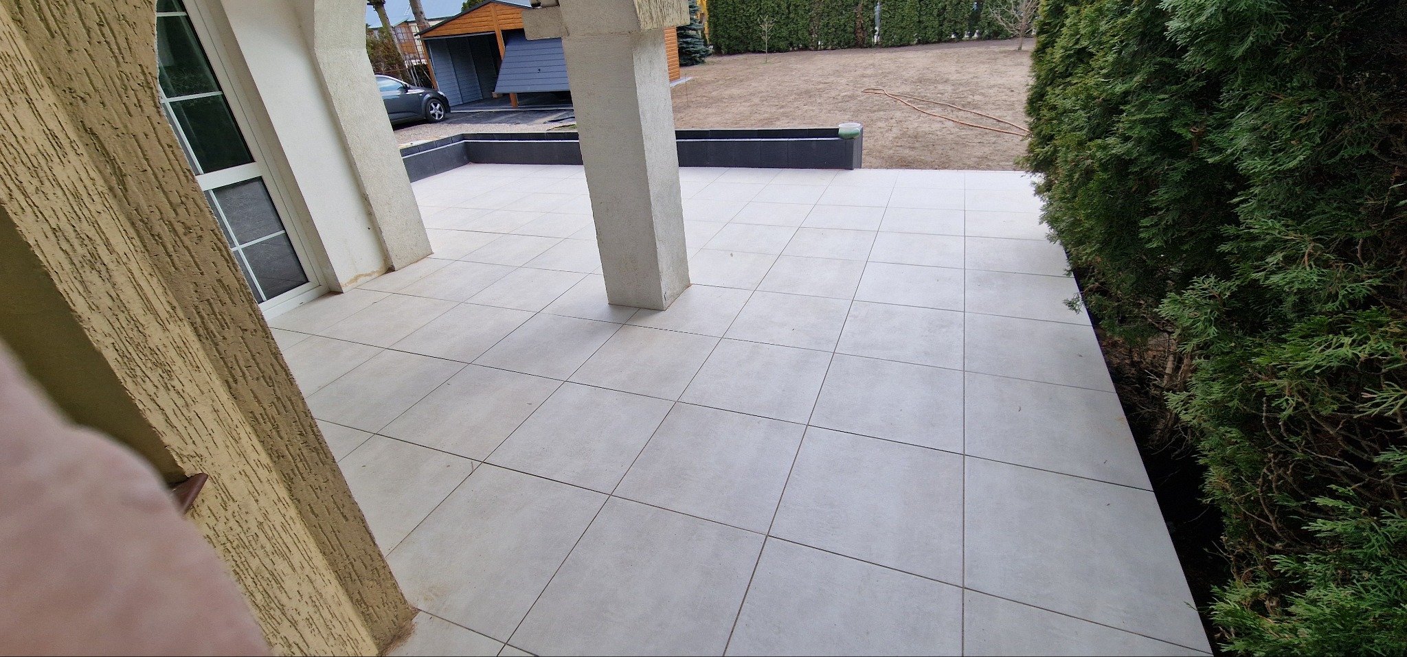 Jasne, kwadratowe płytki tarasowe ułożone na zewnątrz budynku z kolumnami. Widok z góry, zieleń w tle.
