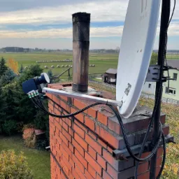 Antena satelitarna zamontowana na kominie z widokiem na wiejski krajobraz z polami i stogami siana w tle.