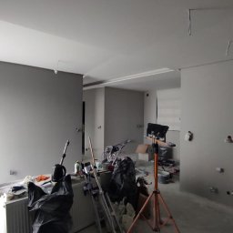 Firma Usługowo-Transportowa Egor Ermolenko - Wnętrze w trakcie prac wykończeniowych: świeżo pomalowane szare ściany, widoczne narzędzia, drabina, lampa budowlana na trójnogu, zakryty grzejnik i worki na śmieci.