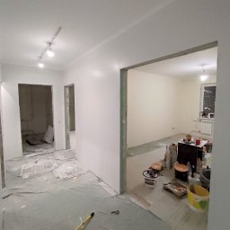Firma Usługowo-Transportowa Egor Ermolenko - Wnętrze mieszkania w trakcie prac wykończeniowych: świeżo przygotowane, białe ściany, zabezpieczona podłoga, widoczne narzędzia i puszki z farbami w sąsiednim pokoju z widokiem na miasto nocą.