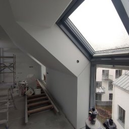 Firma Usługowo-Transportowa Egor Ermolenko - Poddasze w trakcie prac wykończeniowych, widoczne ściany po położeniu gładzi, okno dachowe z widokiem na zewnątrz, drabina i narzędzia malarskie w tle.