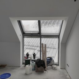 Firma Usługowo-Transportowa Egor Ermolenko - Poddasze po wykonaniu gładzi, widoczne okno dachowe z deszczem, narzędzia i materiały budowlane na podłodze, ściany przygotowane do dalszych prac.