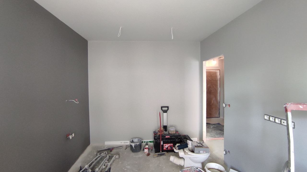 Dwukolorowe wnętrze w trakcie malowania: szara ściana kontrastuje z jasnoszarą, widoczne narzędzia malarskie, instalacja elektryczna i sanitarna w trakcie prac wykończeniowych.