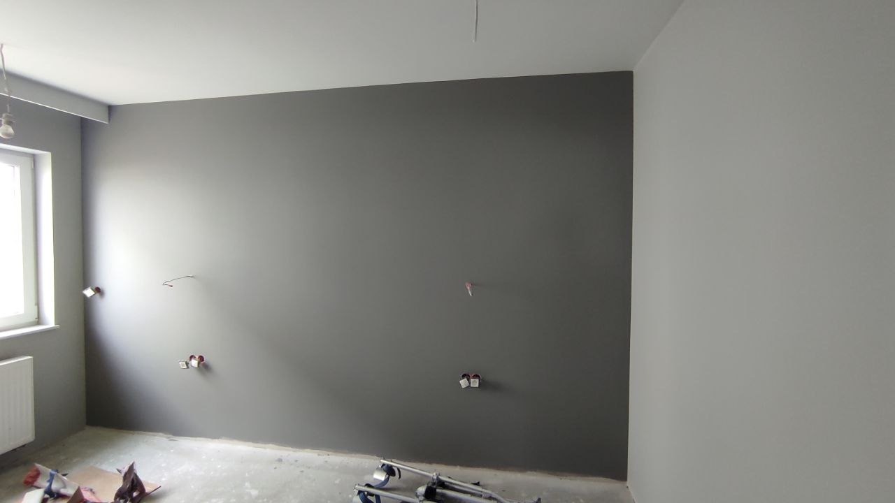 Pokój po świeżym pomalowaniu ścian na dwa odcienie szarości, widoczne puszki elektryczne i wystające kable, na podłodze folia malarska i drabina.