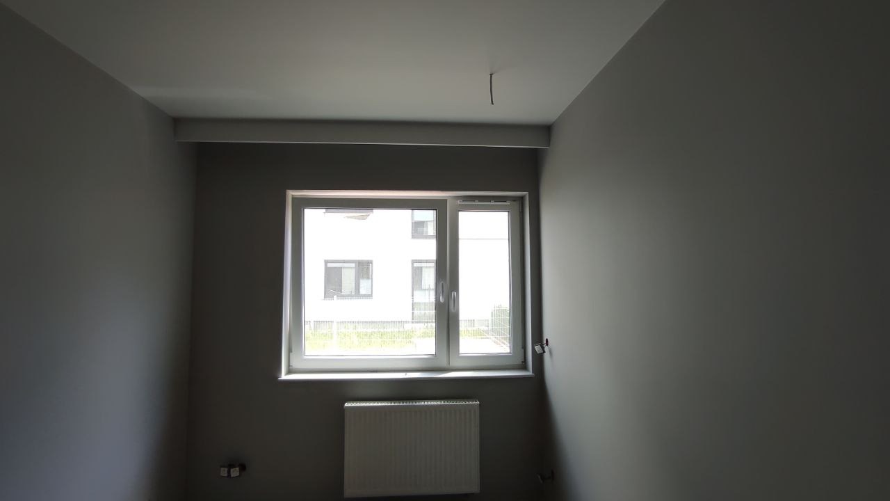 Wnętrze pokoju po remoncie, ściany pomalowane na szaro, białe okno z widokiem na budynek, grzejnik pod oknem, wystający kabel z sufitu.