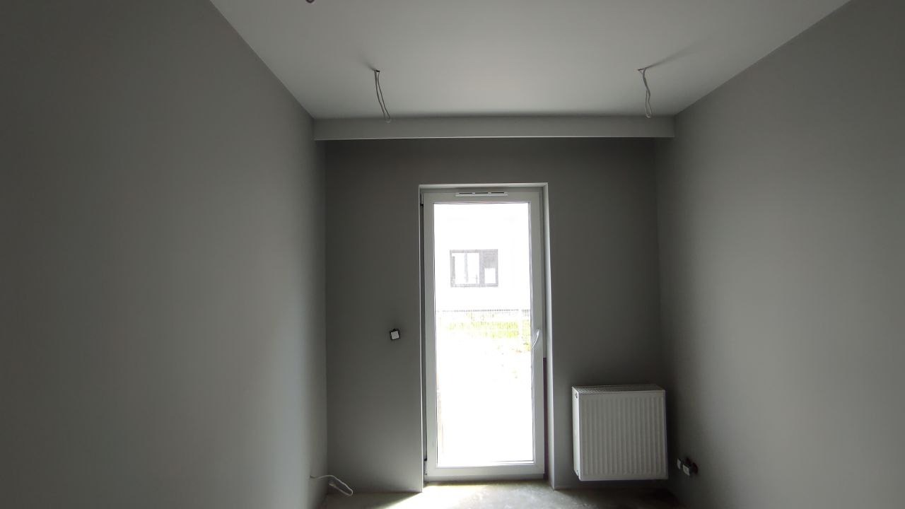Pokój po odświeżeniu, ściany pomalowane na szaro, widoczne kable wystające z sufitu, białe drzwi balkonowe i grzejnik.