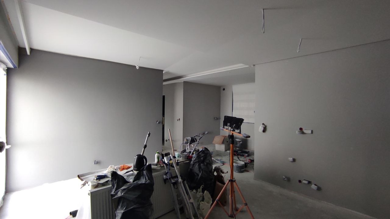 Wnętrze w trakcie prac wykończeniowych: świeżo pomalowane szare ściany, widoczne narzędzia, drabina, lampa budowlana na trójnogu, zakryty grzejnik i worki na śmieci.