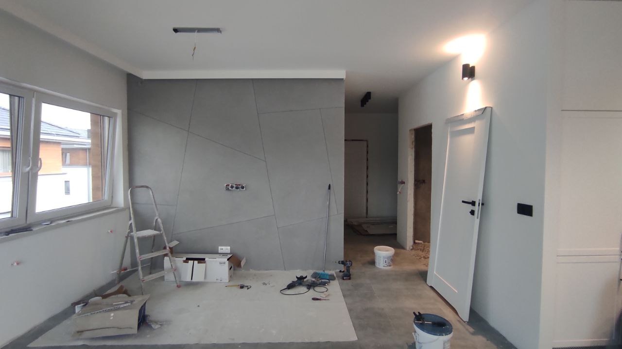 Wnętrze w trakcie prac wykończeniowych: ściana z dekoracyjną strukturą, widoczne narzędzia, drabina, nieotynkowane otwory drzwiowe, surowy beton na podłodze.