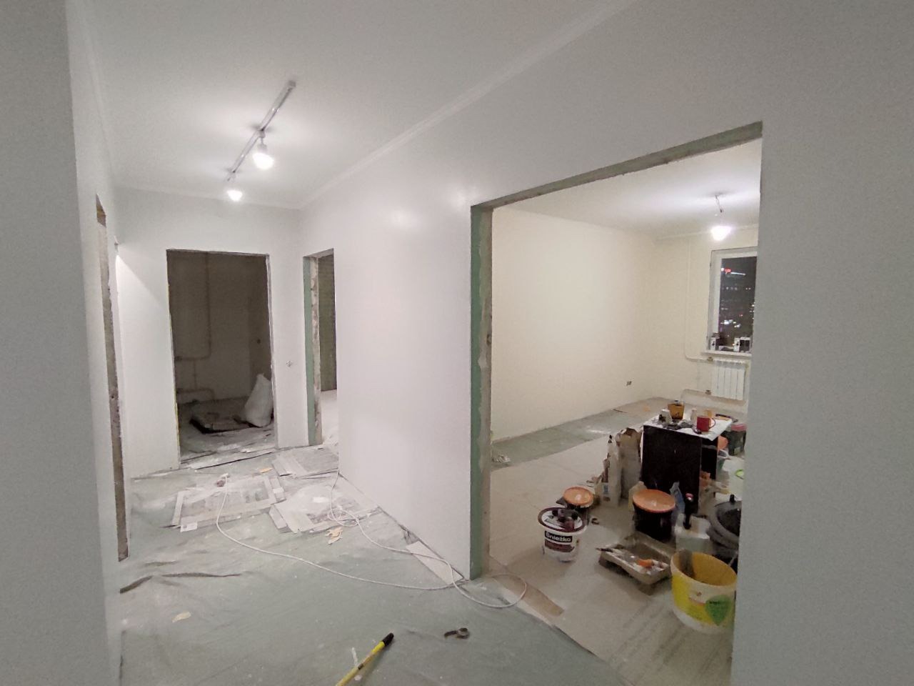 Wnętrze mieszkania w trakcie prac wykończeniowych: świeżo przygotowane, białe ściany, zabezpieczona podłoga, widoczne narzędzia i puszki z farbami w sąsiednim pokoju z widokiem na miasto nocą.