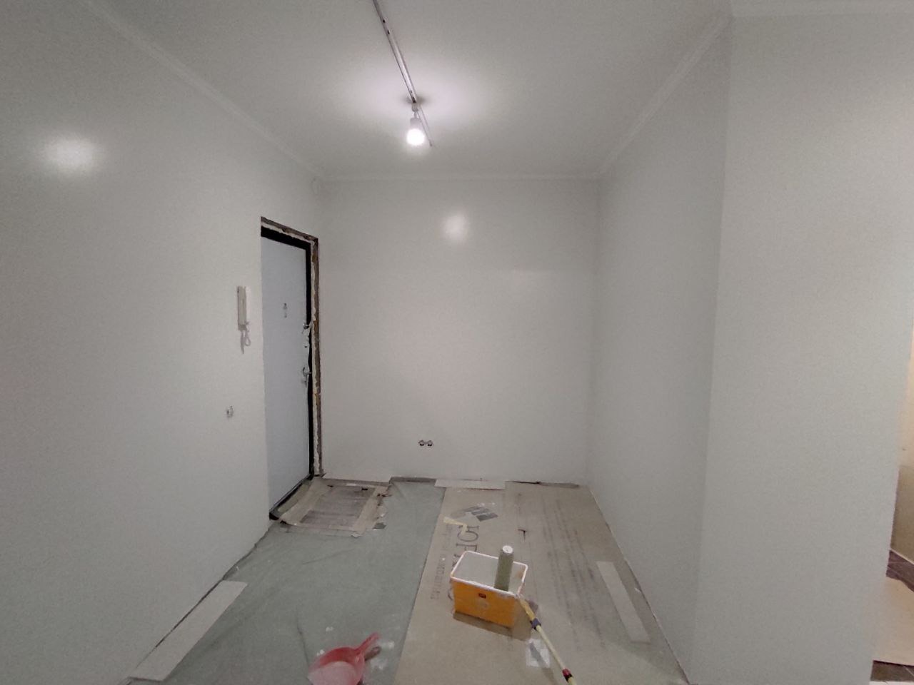 Pomieszczenie po malowaniu na biało, widoczne narzędzia malarskie, zabezpieczona podłoga i ościeżnica drzwiowa.