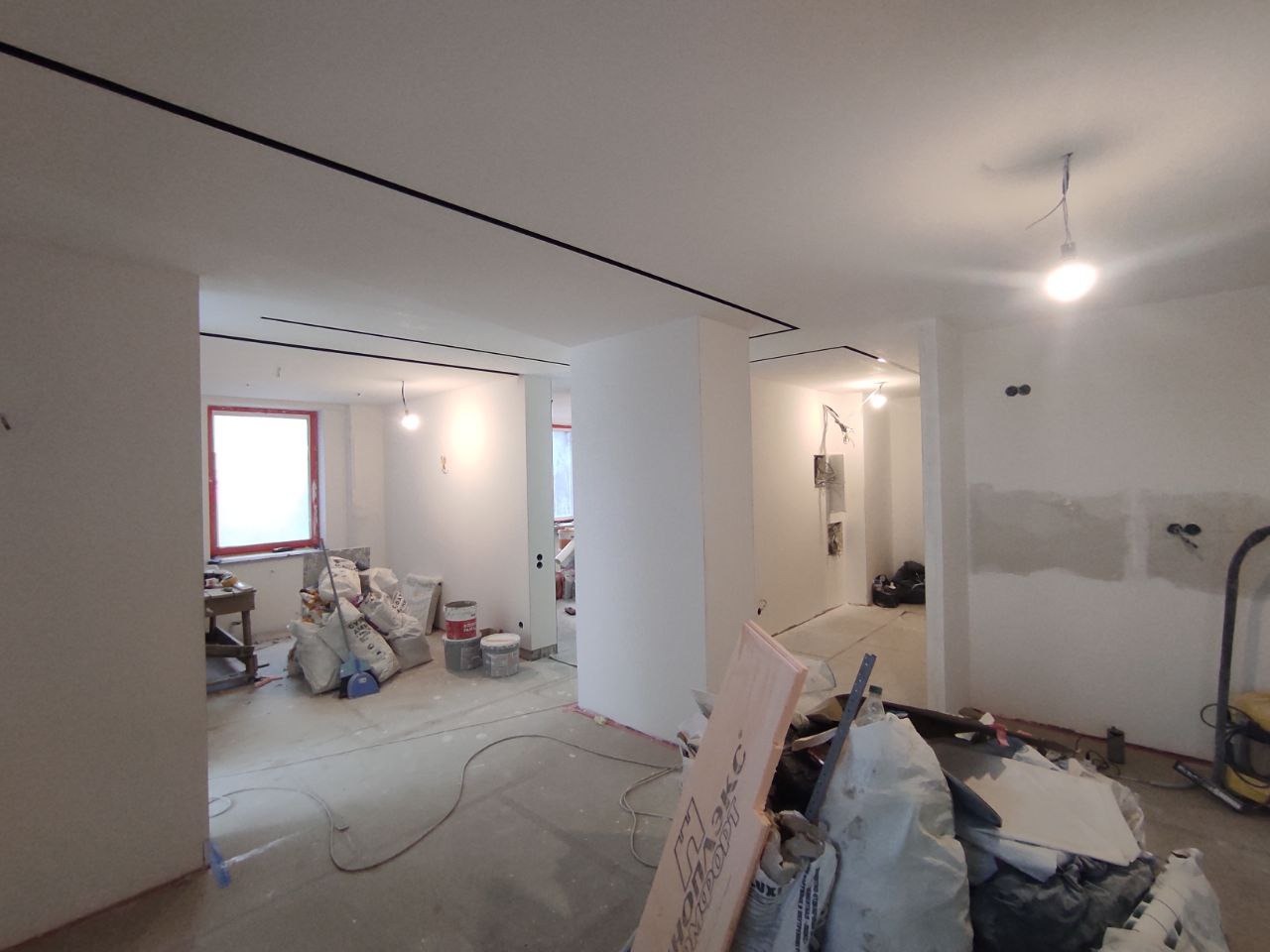 Wnętrze w trakcie prac przygotowawczych: widoczne worki z materiałami budowlanymi, surowe ściany z widocznymi śladami po instalacjach elektrycznych, czarne listwy sufitowe i prowizoryczne oświetlenie.