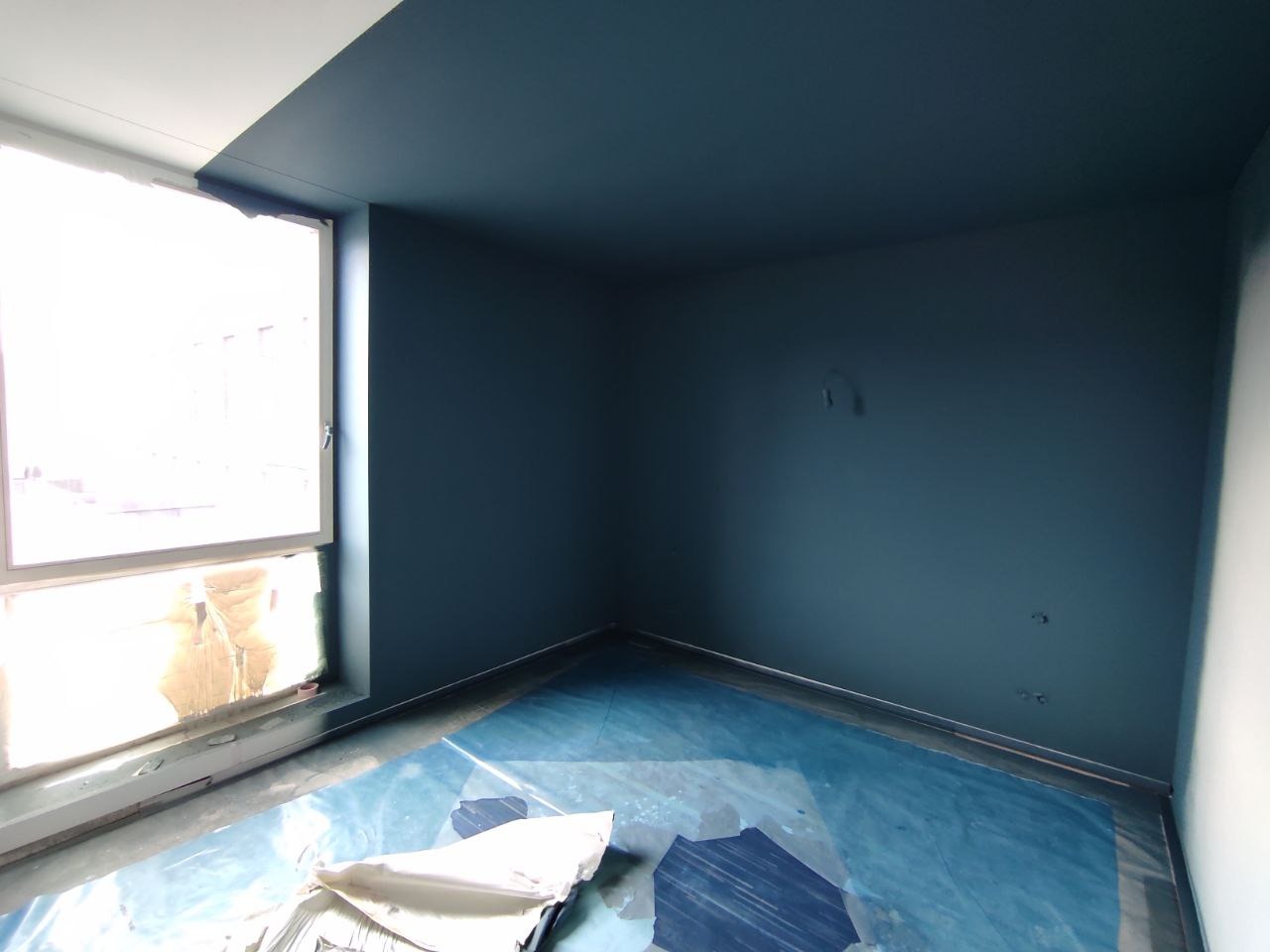 Pomieszczenie po pomalowaniu na ciemny odcień niebieskiego, z zabezpieczoną folią podłogą i oknem zasłoniętym kartonem.