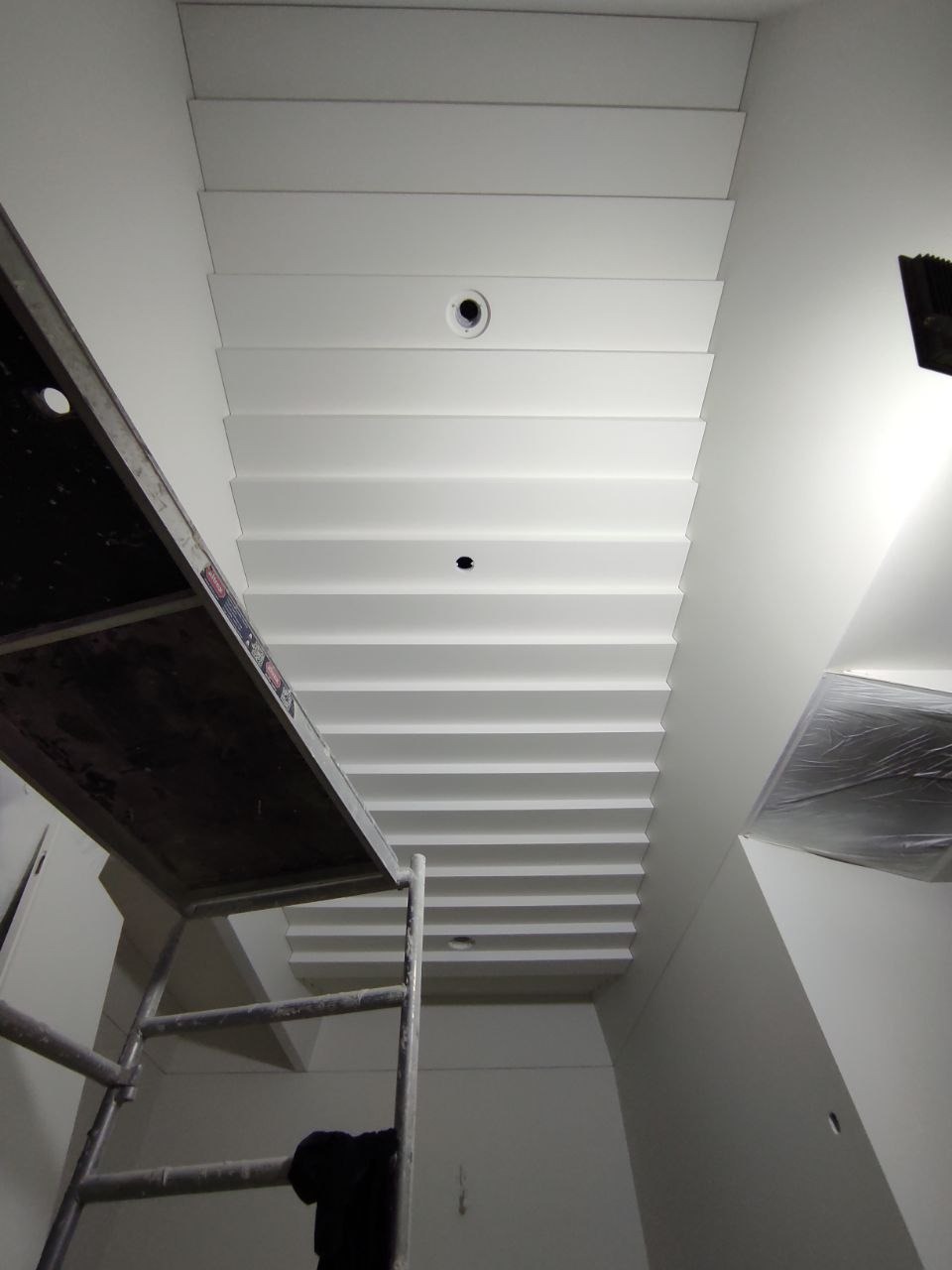 Biały, kasetonowy sufit z oświetleniem punktowym w trakcie prac, widoczny fragment rusztowania i zabezpieczona folią ściana.