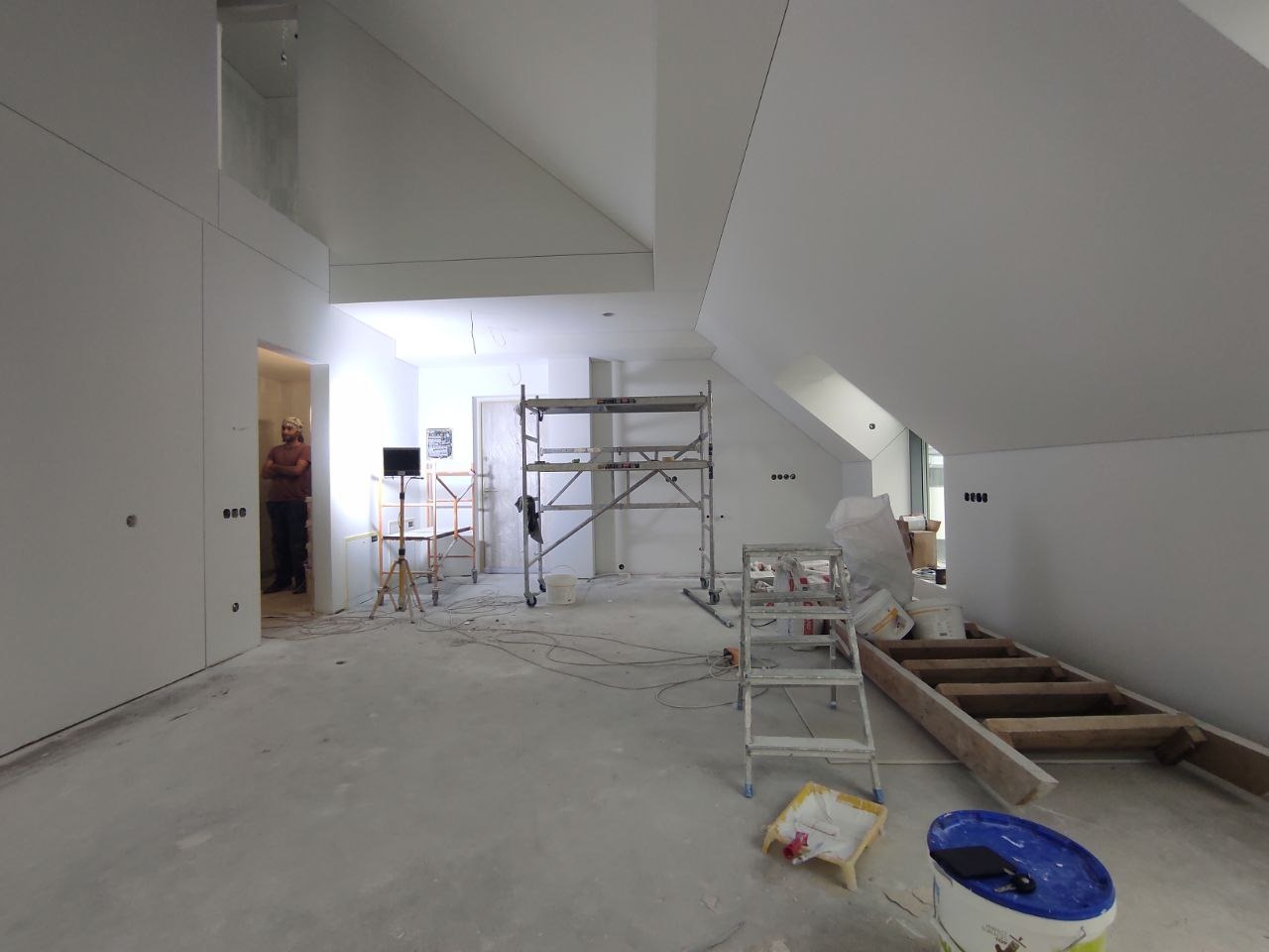 Poddasze w trakcie prac przygotowawczych do dalszego etapu wykończenia, widoczne rusztowania, drabiny, wiadra i narzędzia malarskie, ściany i skosy przygotowane pod dalsze prace wykończeniowe.