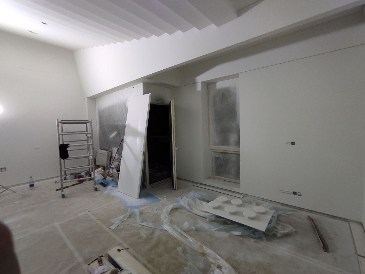 Wnętrze w trakcie prac przygotowawczych, ściany i sufit pomalowane na biało, okno zabezpieczone folią, widoczny drabina i materiały budowlane na podłodze.