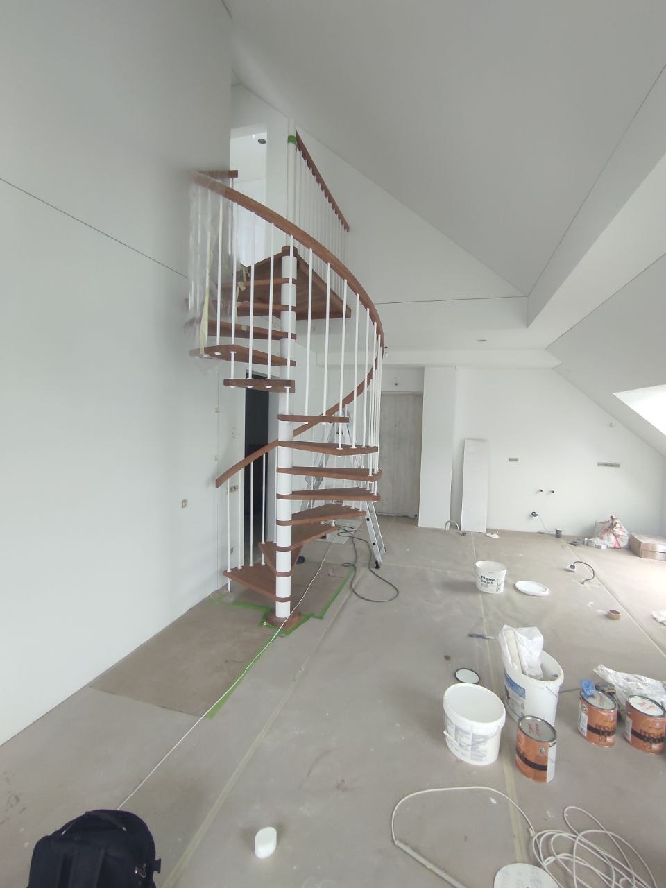 Wnętrze w trakcie prac wykończeniowych: spiralne schody z drewnianymi stopniami i białymi barierkami, zabezpieczone taśmą malarską, widoczne puszki z farbą i drabina w tle.