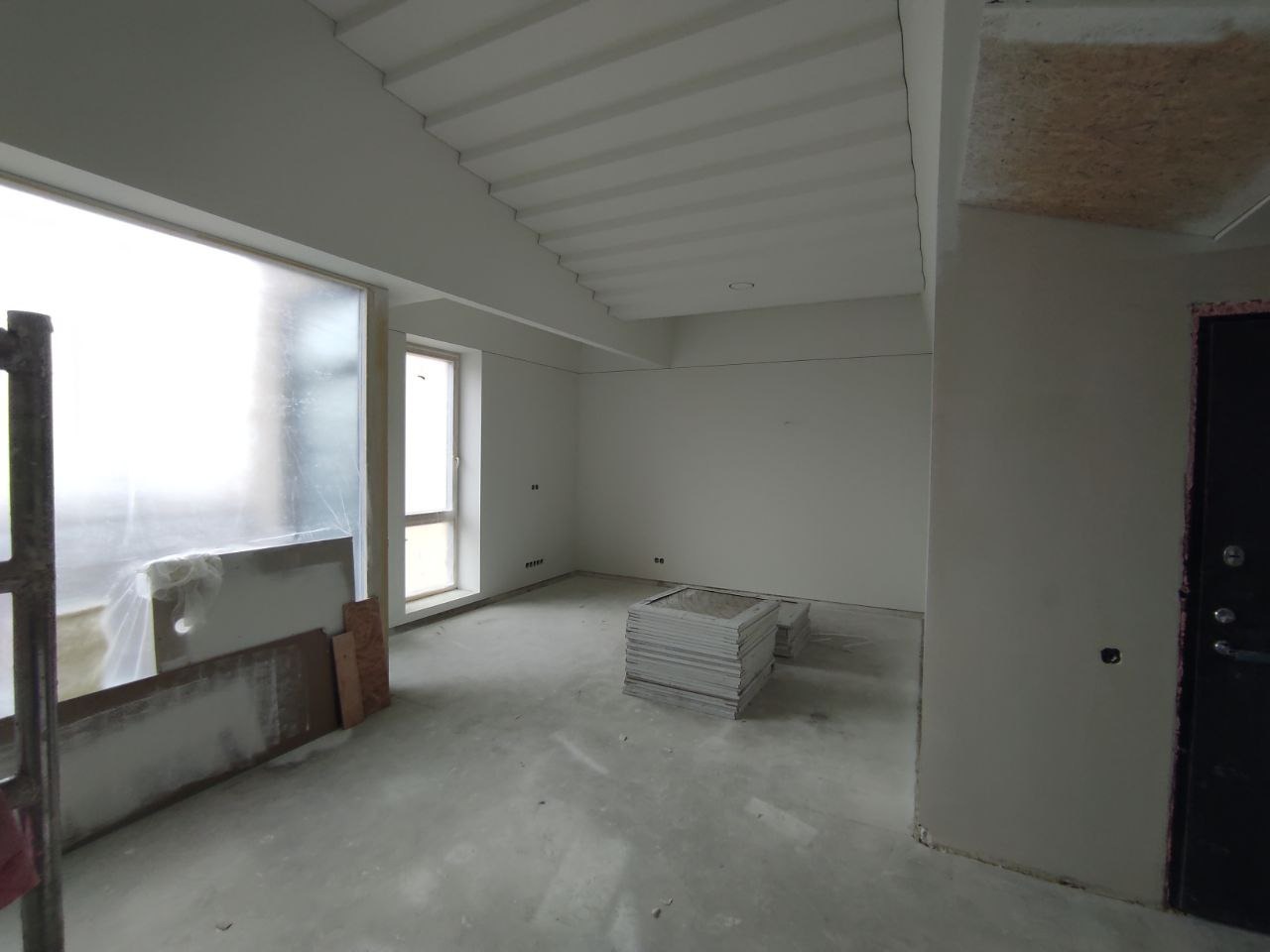 Wnętrze w stanie deweloperskim z widoczną stertą płyt gipsowo-kartonowych, częściowo zakryte okno folią ochronną, sufit z dekoracyjnymi listwami.