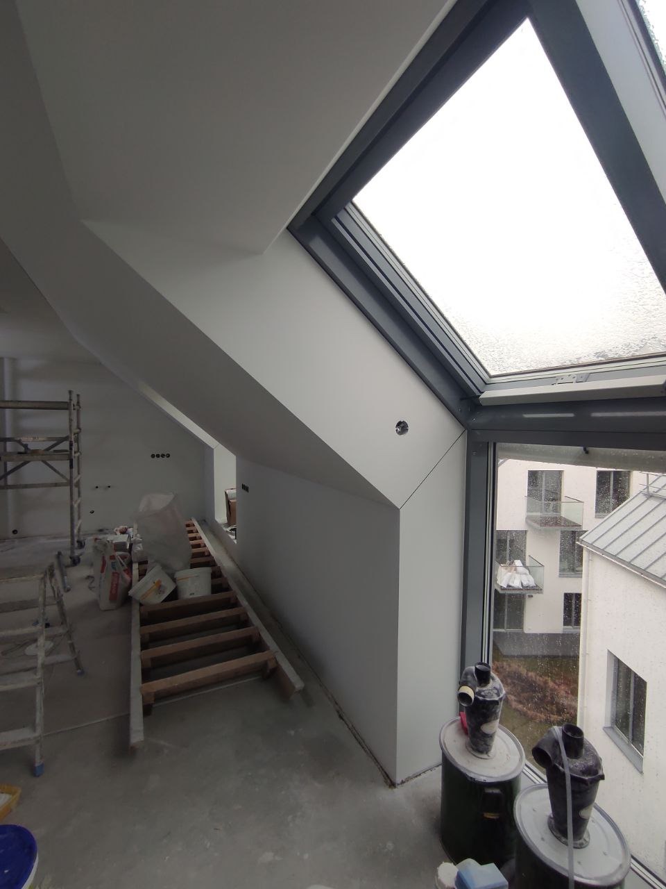 Poddasze w trakcie prac wykończeniowych, widoczne ściany po położeniu gładzi, okno dachowe z widokiem na zewnątrz, drabina i narzędzia malarskie w tle.