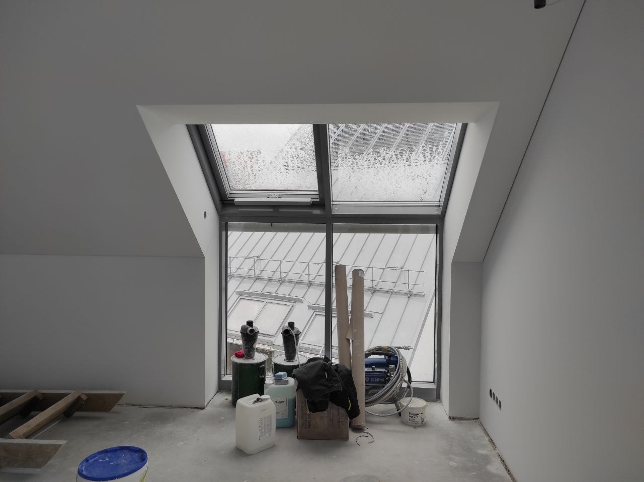 Poddasze po wykonaniu gładzi, widoczne okno dachowe z deszczem, narzędzia i materiały budowlane na podłodze, ściany przygotowane do dalszych prac.